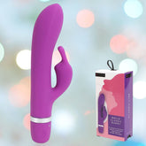 A B Swish Bwild Classic G-Spot and Clitoral Vibrator in purple is displayed next to its packaging against a soft, blurred background with pastel circles of light. The waterproof product features a main shaft and a smaller external stimulator, with the label "Bwild Classic Bunny" visible on the box.